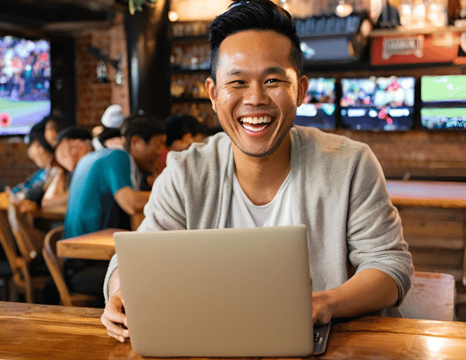 An Indonesian man, with his laptop open on a wooden table, smiling brightly in a well-lit sports bar filled with sports fans and large screens.
